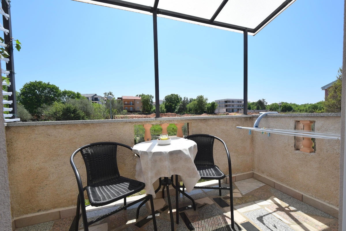 A small balcony is presented, featuring a round table set with a white tablecloth and two black chairs. A scenic view of lush green areas and buildings is visible in the background. Bright blue skies create a serene atmosphere.