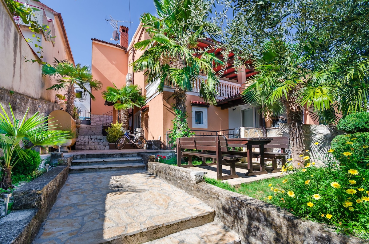 A charming outdoor area features stone pathways leading to a two-story building surrounded by lush greenery and palm trees. A wooden dining table is positioned on a stone terrace, offering a serene space to relax. Vibrant flowers add color to the landscape.
