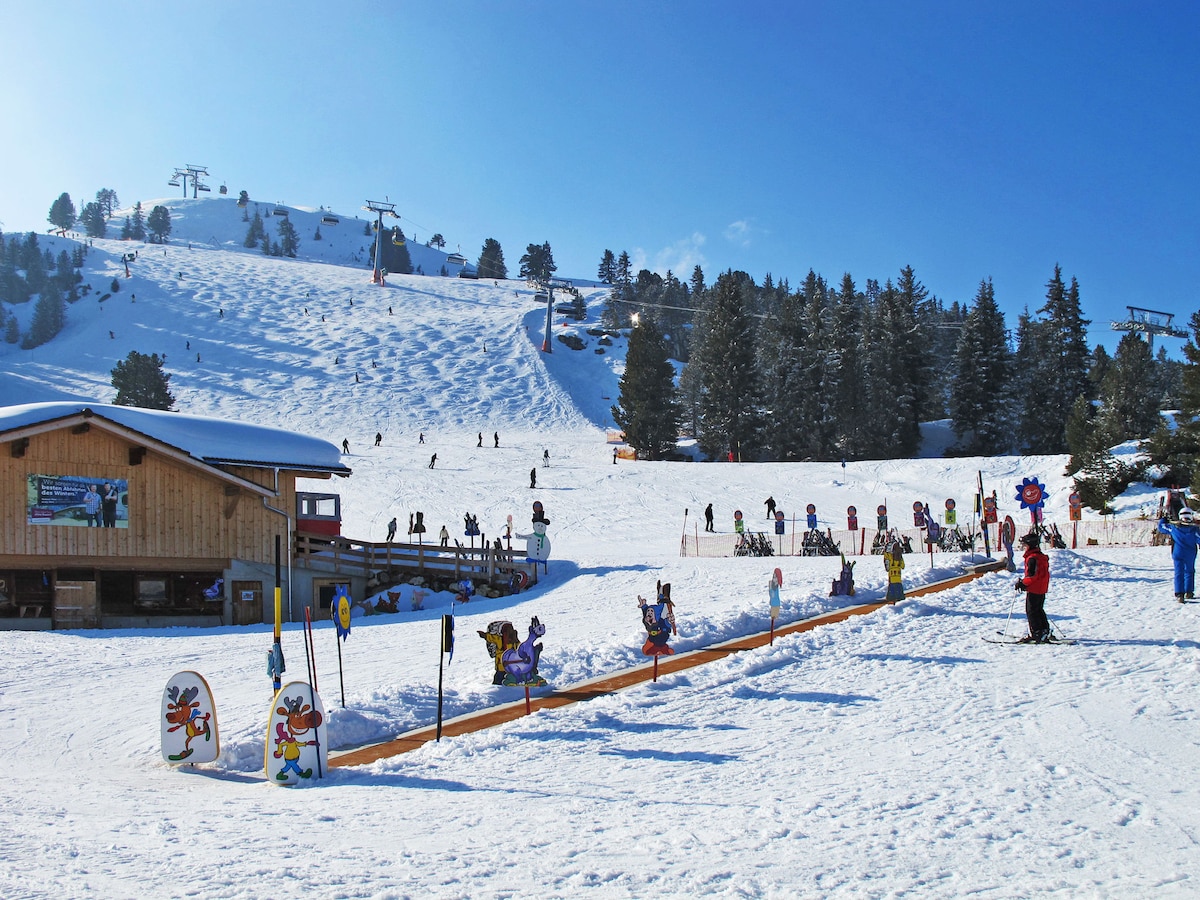 A winter scene captures a snow-covered hill with ski lifts in the distance. Children and adults are skiing and playing in the snow. Colorful signage decorates the pathway in the foreground, providing a playful contrast to the white landscape and evergreen trees lining the area.