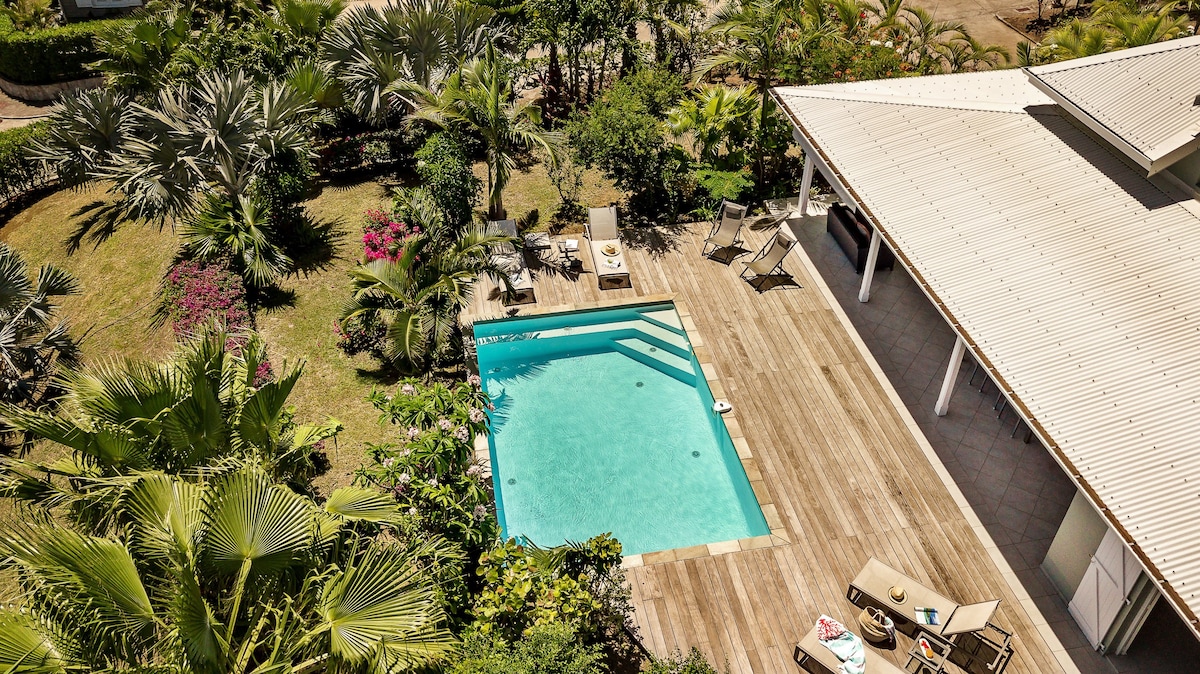 An aerial view of the villa's private swimming pool is shown, surrounded by a wooden deck and lush tropical foliage. Sun loungers are arranged around the pool, inviting relaxation. The vibrant greenery and colorful flowers enhance the outdoor ambiance.