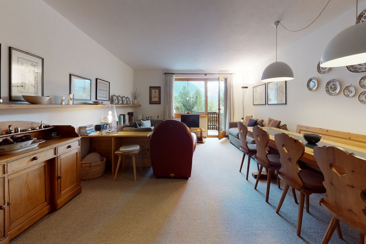 The spacious living and dining area features a wooden dining table surrounded by several chairs. A lounge area with a sofa and armchair is positioned near a large window, offering natural light and access to the terrace. Decorative plates and framed artwork adorn the walls.