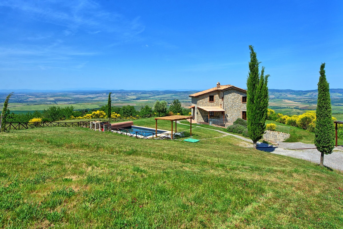 A rustic villa sits atop a small hill, surrounded by lush greenery with expansive views of the Val d'Orcia valleys. A private swimming pool and terrace area can be seen, complemented by neatly arranged sun loungers and shade structures.