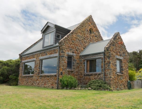 The rustic stone cottage is showcased from an exterior angle, highlighting its unique stone facade and steeply pitched roof. Large windows invite natural light, while the surrounding grassy area features low shrubbery and a vibrant backdrop of trees, contributing to the secluded environment.