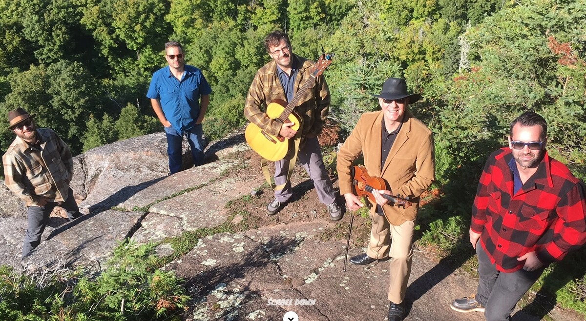 A group of five individuals is positioned on a rocky outcrop surrounded by lush greenery. One person is playing an acoustic guitar, while another holds a violin. The natural landscape offers a serene setting, contributing to a sense of togetherness in a scenic environment.