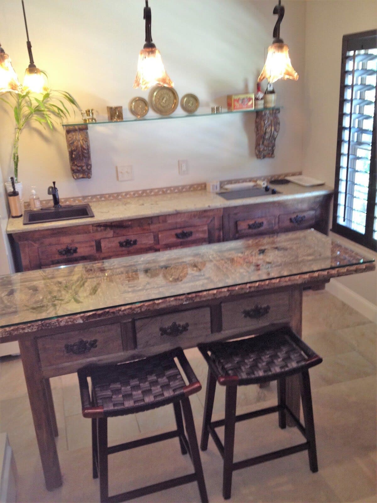 The kitchenette features a wooden countertop with a glossy finish and two woven stools. Upper shelves display decorative plates and small items. A double sink and modern faucet are included, with an additional countertop area for food preparation, providing functionality in a stylish setting.