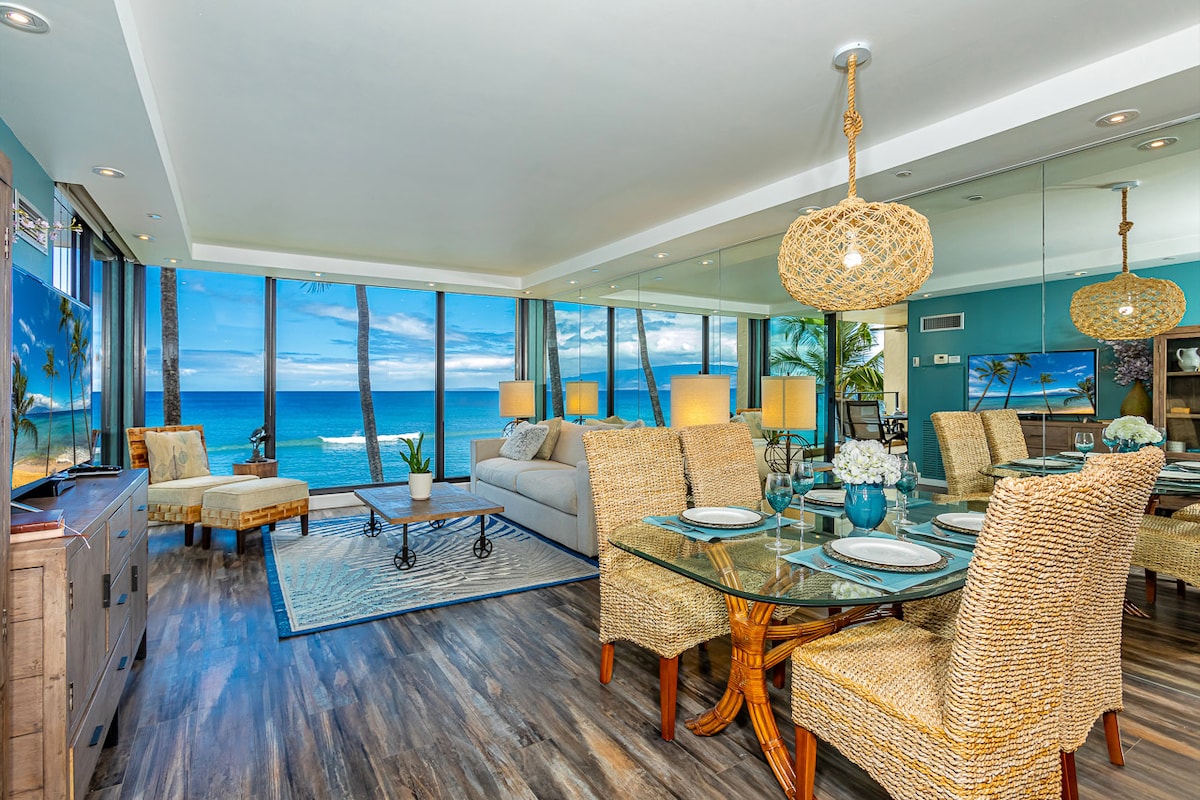 A bright living area features large windows showcasing ocean views, allowing natural light to illuminate the space. A comfortable seating arrangement includes a light-colored sofa and a glass dining table surrounded by woven chairs. Coastal decor accents enhance the serene atmosphere.