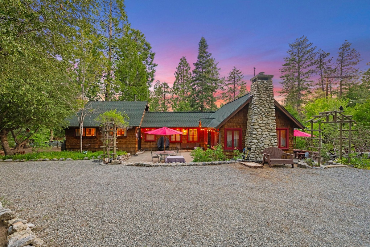 A charming cabin is surrounded by trees, with a stone chimney and a cozy front patio. Red umbrellas offer shade over outdoor seating areas, while a gravel pathway leads to the entrance. The evening sky is illuminated in soft pink and purple hues, enhancing the tranquil atmosphere.