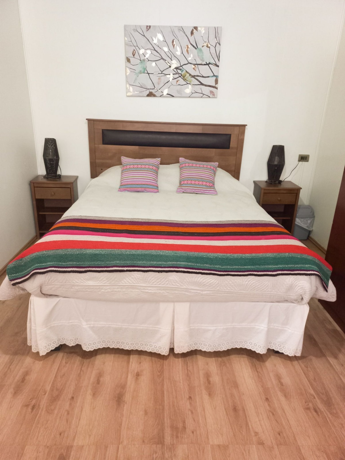 A spacious bed is presented with a colorful striped blanket and two accent pillows. Nightstands with lamps are placed on either side of the bed. A piece of wall art featuring abstract designs is displayed above the headboard, adding a touch of decor to the room.