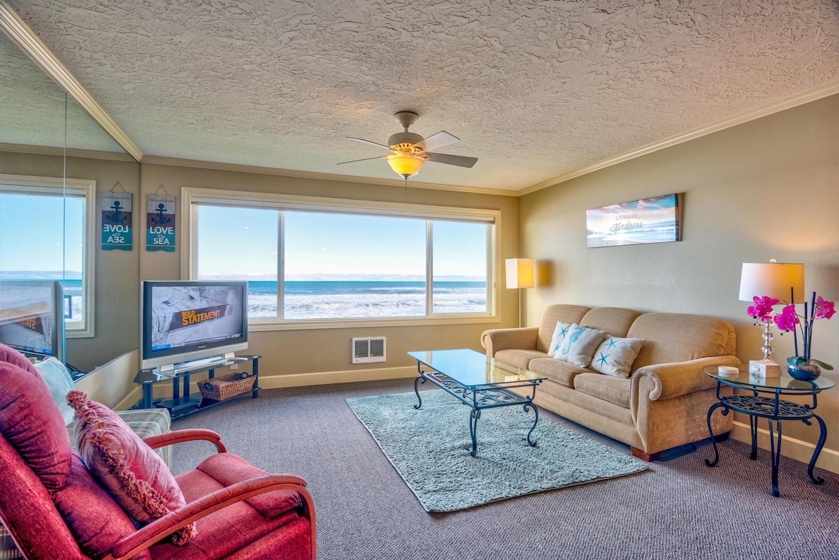 A cozy living area is presented with soft seating and a glass coffee table. Large windows provide a view of the ocean, while a ceiling fan and light fixtures enhance the environment. A television stands on a media console, complemented by decorative elements.