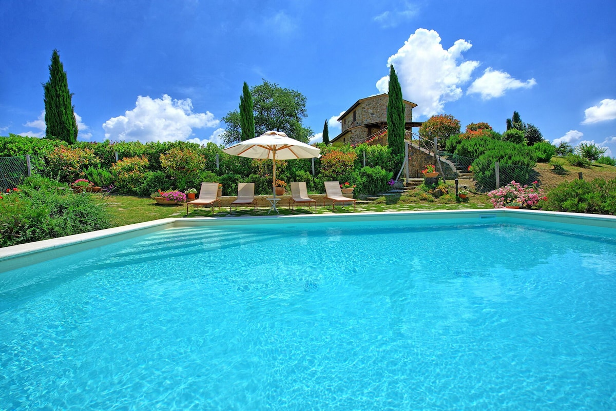 A private swimming pool is surrounded by a well-maintained garden featuring vibrant flowers and greenery. Comfortable sun loungers are positioned beside the pool under a large umbrella, providing shade. The charming stone barn in the background complements the serene landscape under a clear blue sky.