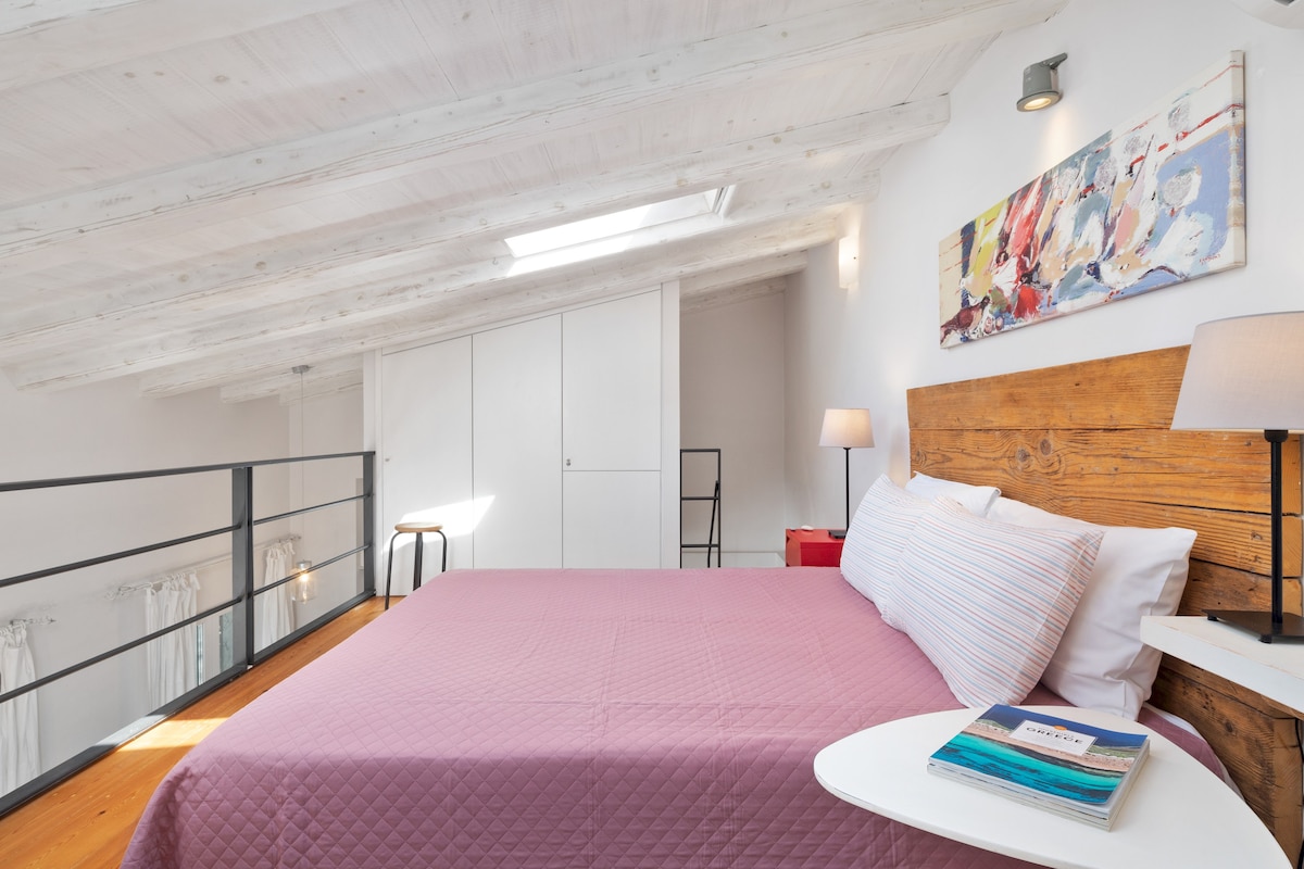 A cozy sleeping area features a snug double bed covered with a soft pink quilt. Natural light enters through a skylight and nearby windows, illuminating the rustic wooden wall and accentuating the simplicity of the design. A bedside table holds a stack of magazines.