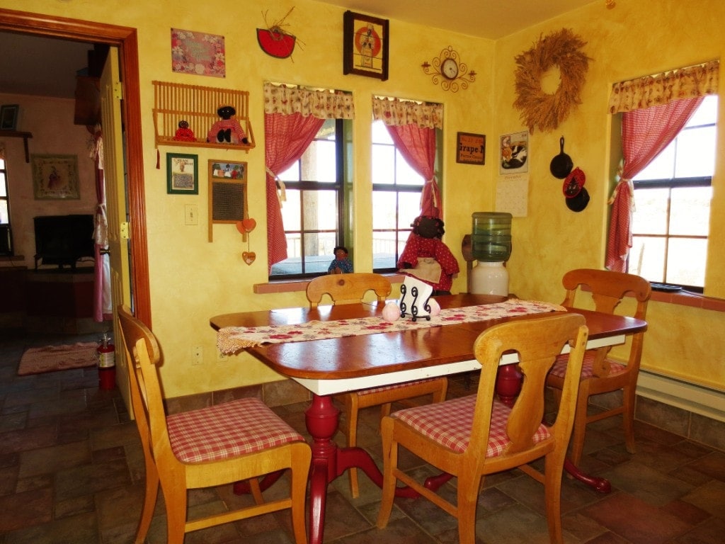 A dining area features a wooden table with a red and white checkered table runner, surrounded by four wooden chairs. Brightly colored walls are adorned with various decorative items and window treatments, creating a cheerful atmosphere. A water cooler stands beside one window that offers natural light.