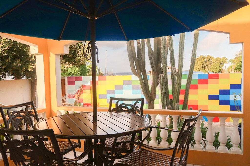 A shaded outdoor dining area features a round table with six matching chairs, surrounded by vibrant garden views. A large umbrella provides shade, while a colorful mural and cacti are visible in the background, creating a lively and inviting atmosphere.