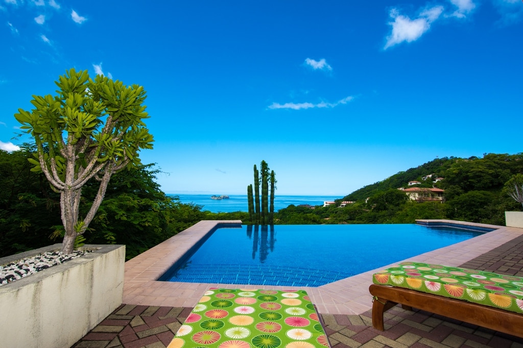 A tranquil infinity pool reflects the vibrant blue sky and ocean view. Lush greenery frames the scene, with a palm tree visible near the pool's edge. Colorful lounge chairs are positioned for relaxation, inviting guests to enjoy the serene surroundings.