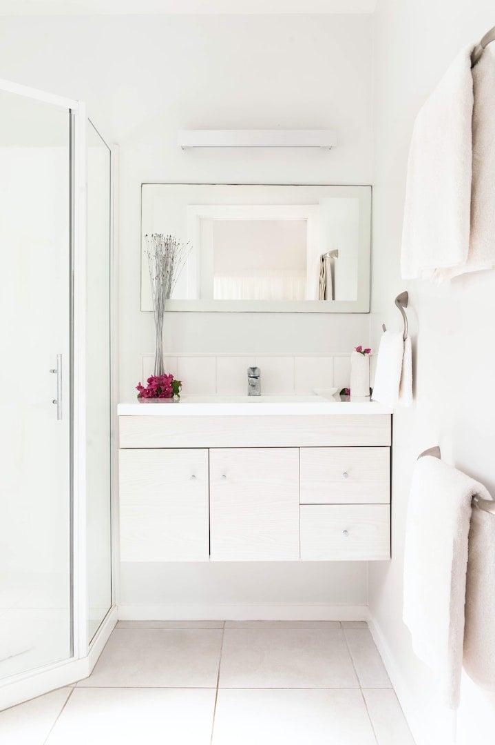 A bright and modern bathroom features a glass shower and a stylish vanity with four drawers. A mirror above the sink reflects the white walls, while soft towels hang on the side. A small vase with flowers adds a touch of color.