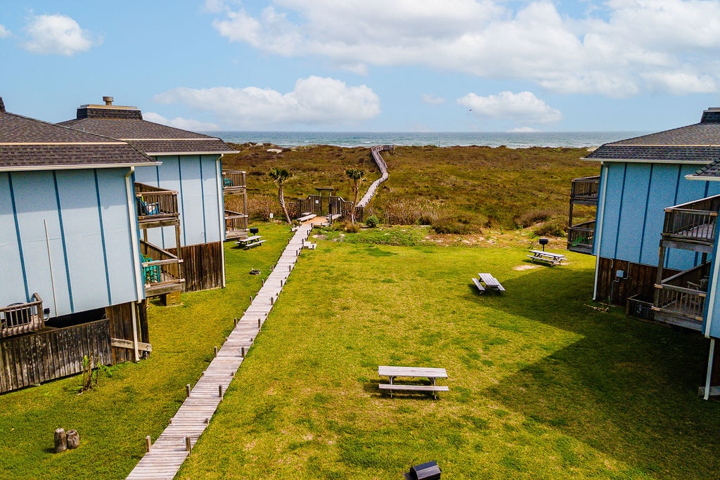 A view of the outdoor space showcases a well-maintained grassy area with picnic tables and a boardwalk leading towards the beach. Surrounding buildings exhibit a coastal design, complemented by a backdrop of blue skies and distant ocean views.