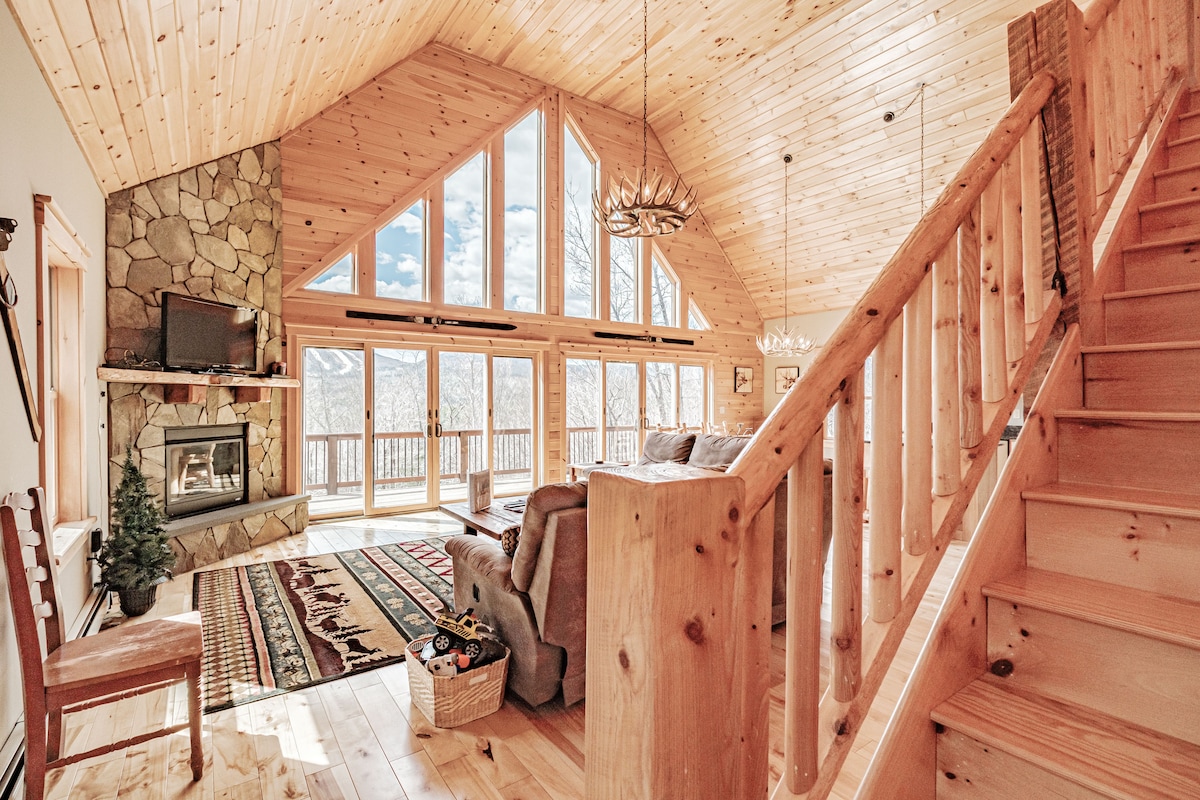 A spacious living area features warm wood finishes, high cathedral ceilings, and large windows offering expansive mountain views. A stone fireplace anchors the room, while a soft seating arrangement invites relaxation. Stairs lead to the upper level, adding architectural interest to the open layout.