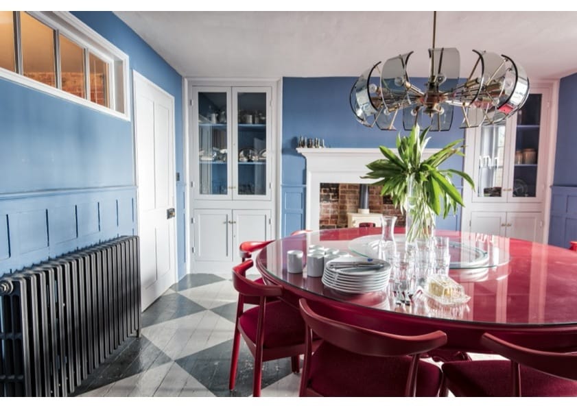 A vibrant dining room features a circular red table surrounded by matching chairs. A sleek chandelier hangs above, illuminating the space. Built-in cabinets display dishware, and a cozy fireplace adds character, all complemented by the fresh green centerpiece.