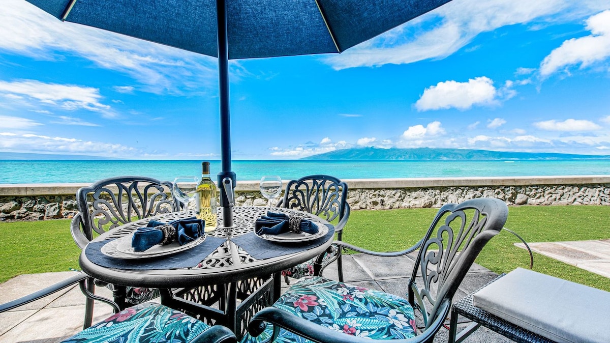 A round outdoor dining set with floral-patterned cushions is shaded by a blue umbrella. The table is laid with plates and glasses, offering a view of the ocean and nearby islands. The vibrant sky and turquoise waters provide a bright backdrop.