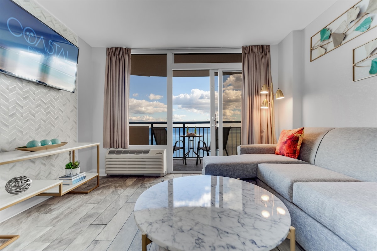 A modern living area is depicted, featuring a comfortable gray sofa and a round marble coffee table. A flat-screen television is mounted on the wall. Floor-to-ceiling glass doors offer a view of the ocean, leading to a small balcony with outdoor seating.