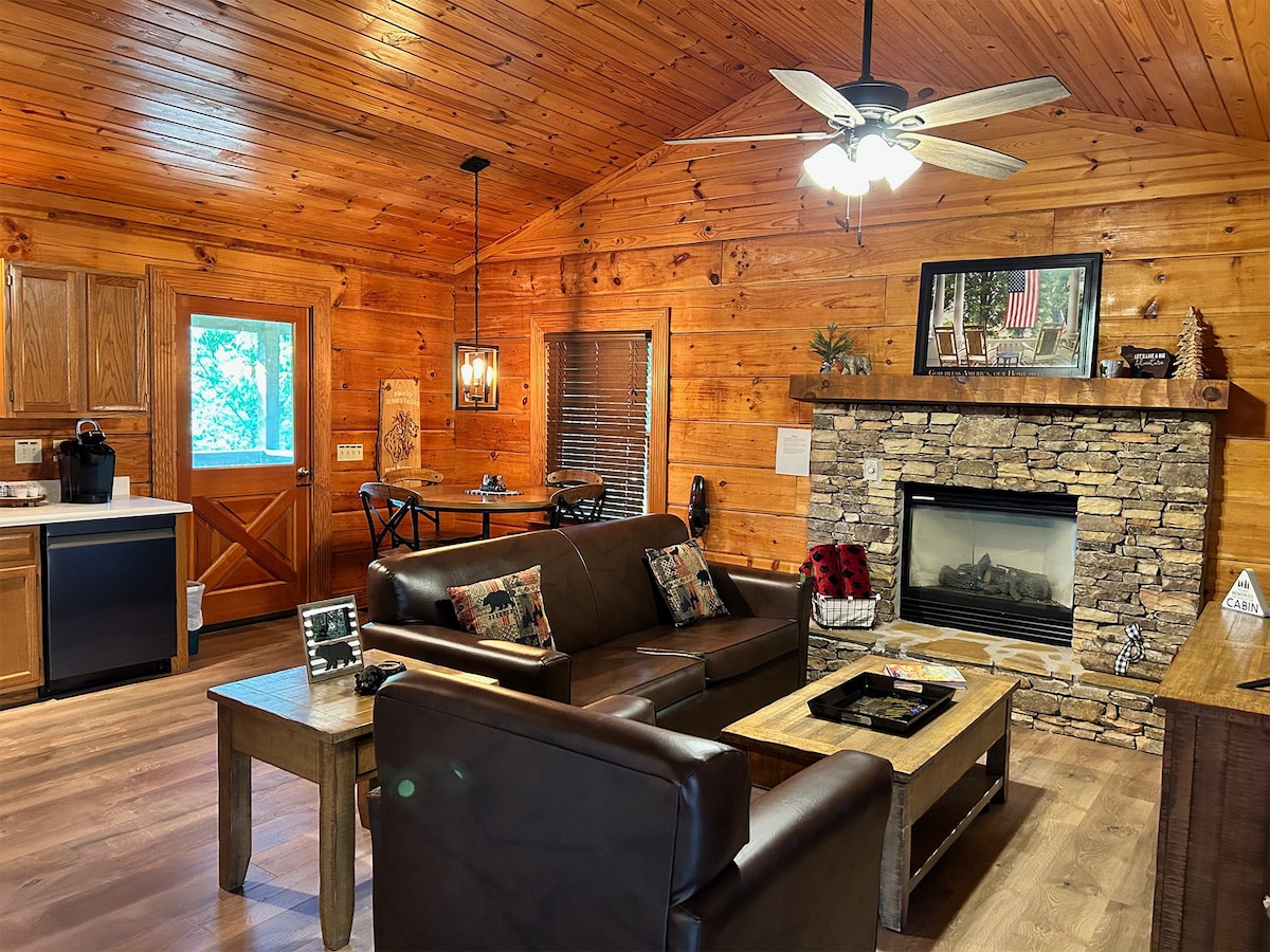 A spacious living area features wooden walls and ceiling, enhancing the cabin's rustic charm. A comfortable dark leather sofa is centered in front of a stone fireplace, with a wooden coffee table nearby. Appliances and a dining table are visible, providing convenient functionality.