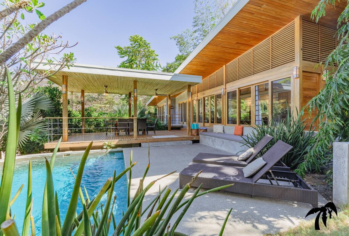 A modern home with a spacious outdoor area is showcased, featuring a bright poolside deck. Sun loungers are positioned near the refreshing pool, surrounded by tropical landscaping and large sliding glass doors that invite natural light into the living space.