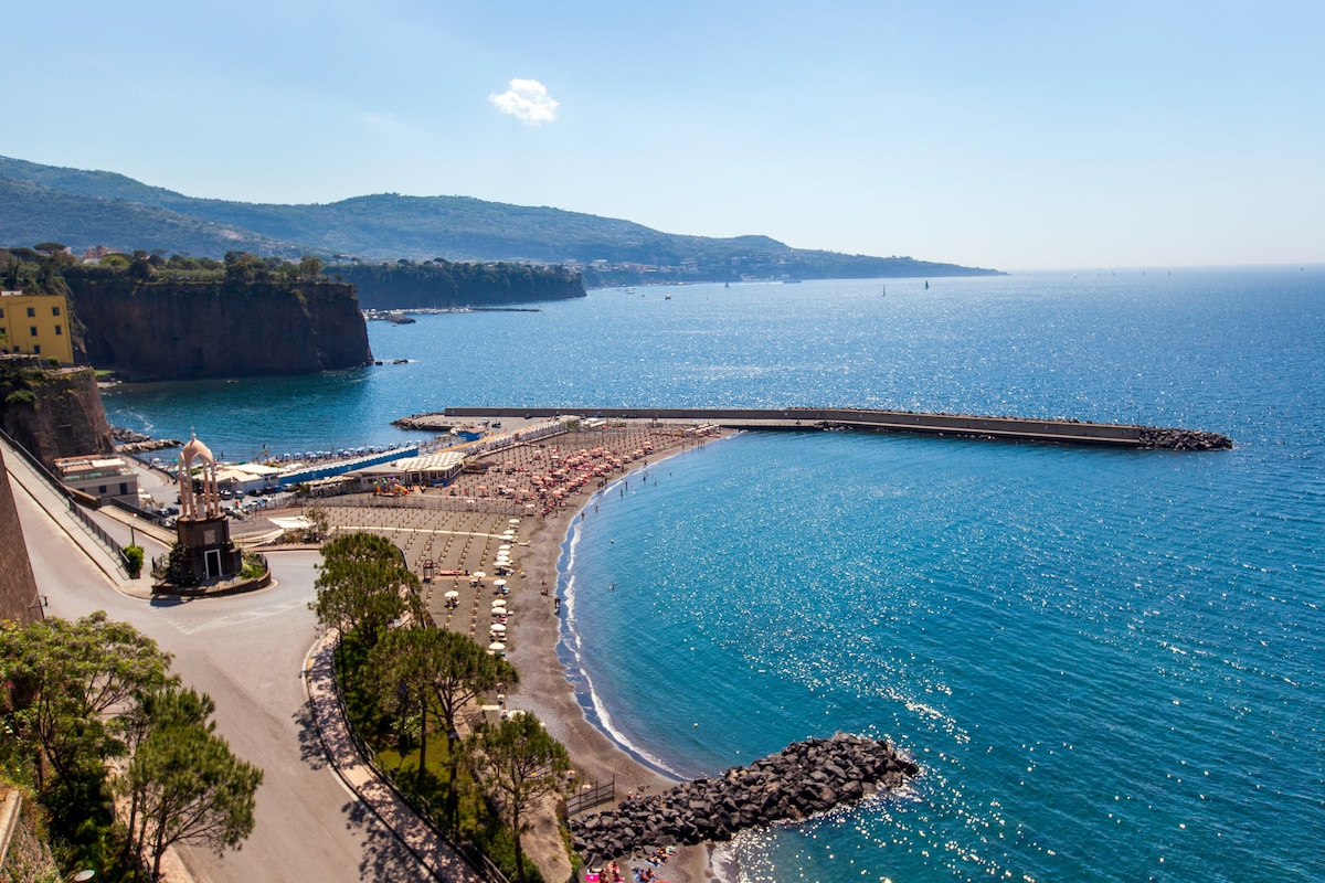 A panoramic view of the coastline showcases a serene stretch of blue water meeting a sandy beach. Sun loungers are lined along the shore, and a curved pier extends into the sea, framed by lush hills in the background under a clear sky.