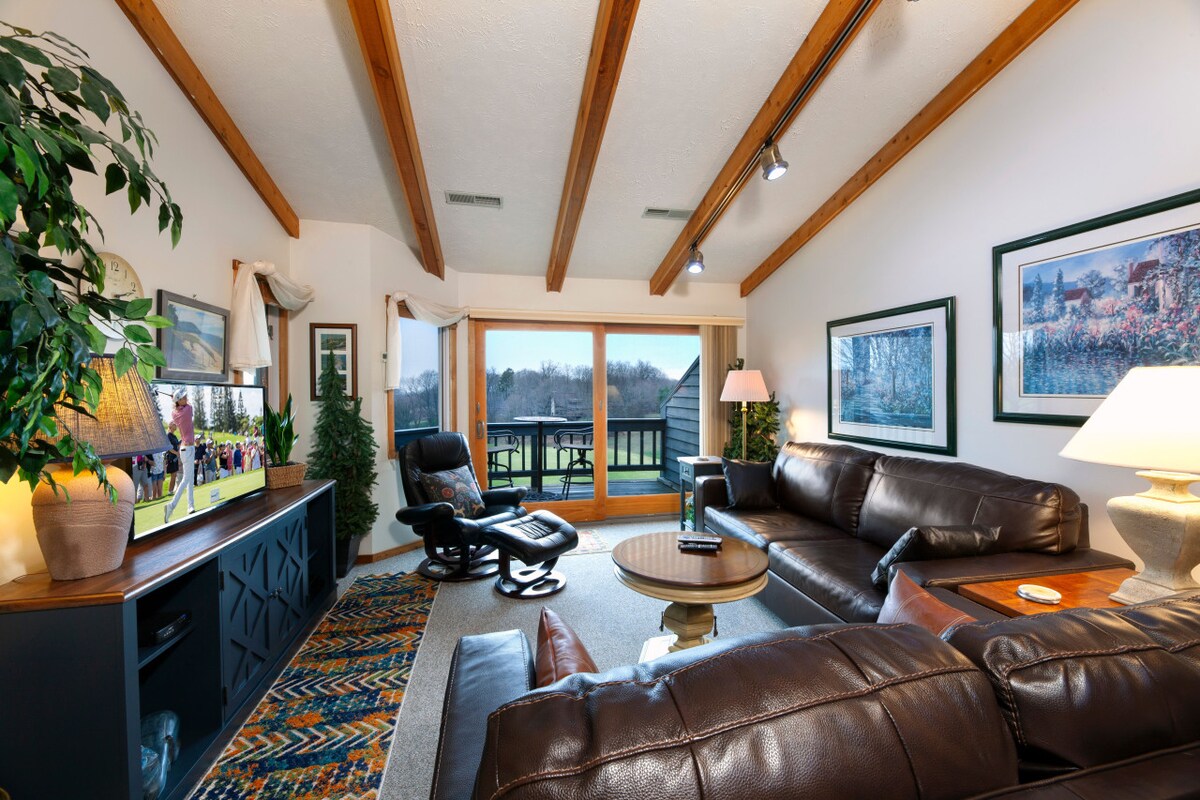 A spacious living area is highlighted by vaulted wooden beams and large windows offering views of the outdoor space. Comfortable leather furniture surrounds a coffee table, while a decorative area rug adds color. A TV stand and potted plants complete the inviting atmosphere.