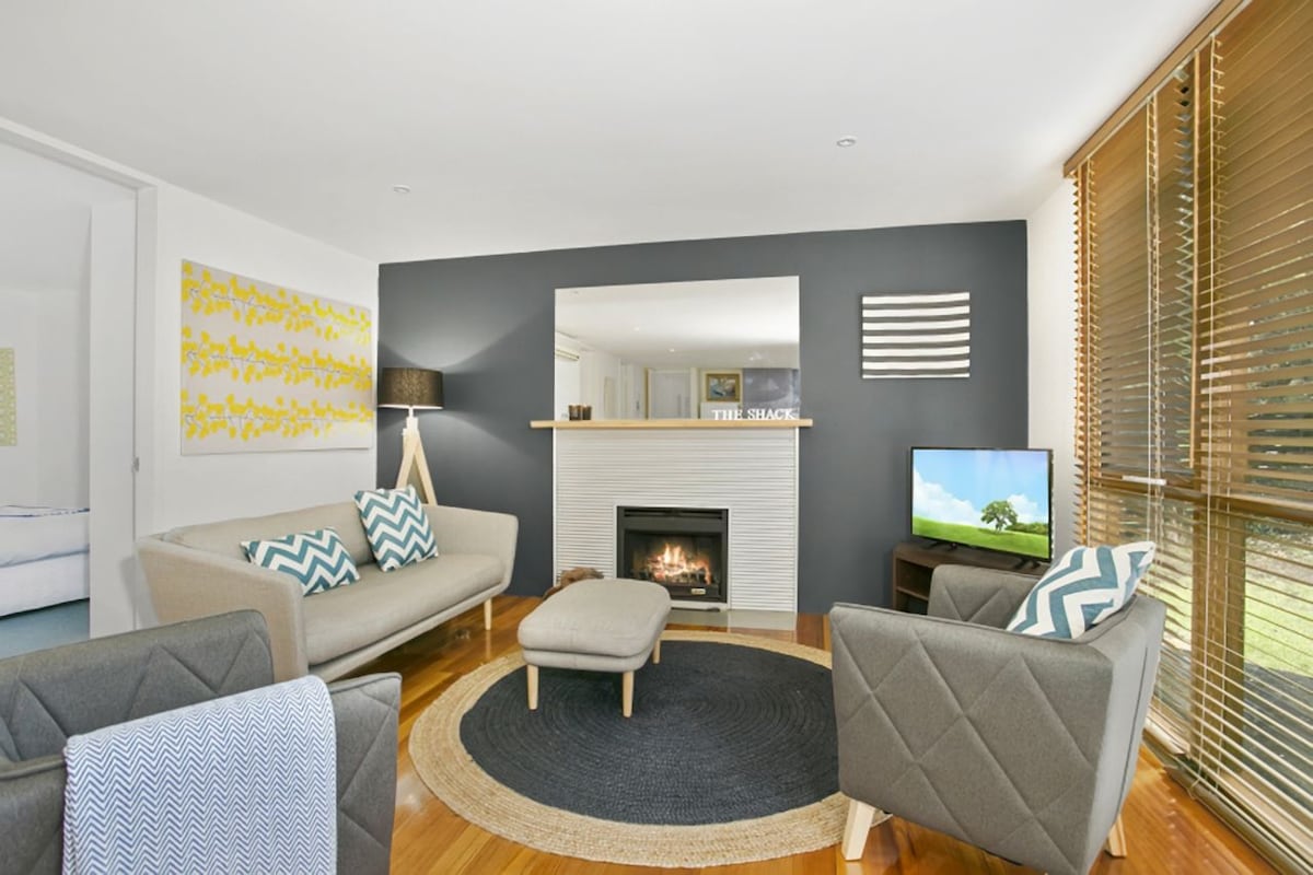 A cozy living area features two light gray sofas and a matching ottoman set on a circular rug. A fireplace is set within a white wall, while a small television sits on a stand. Natural light filters through wooden blinds, creating a warm ambiance.