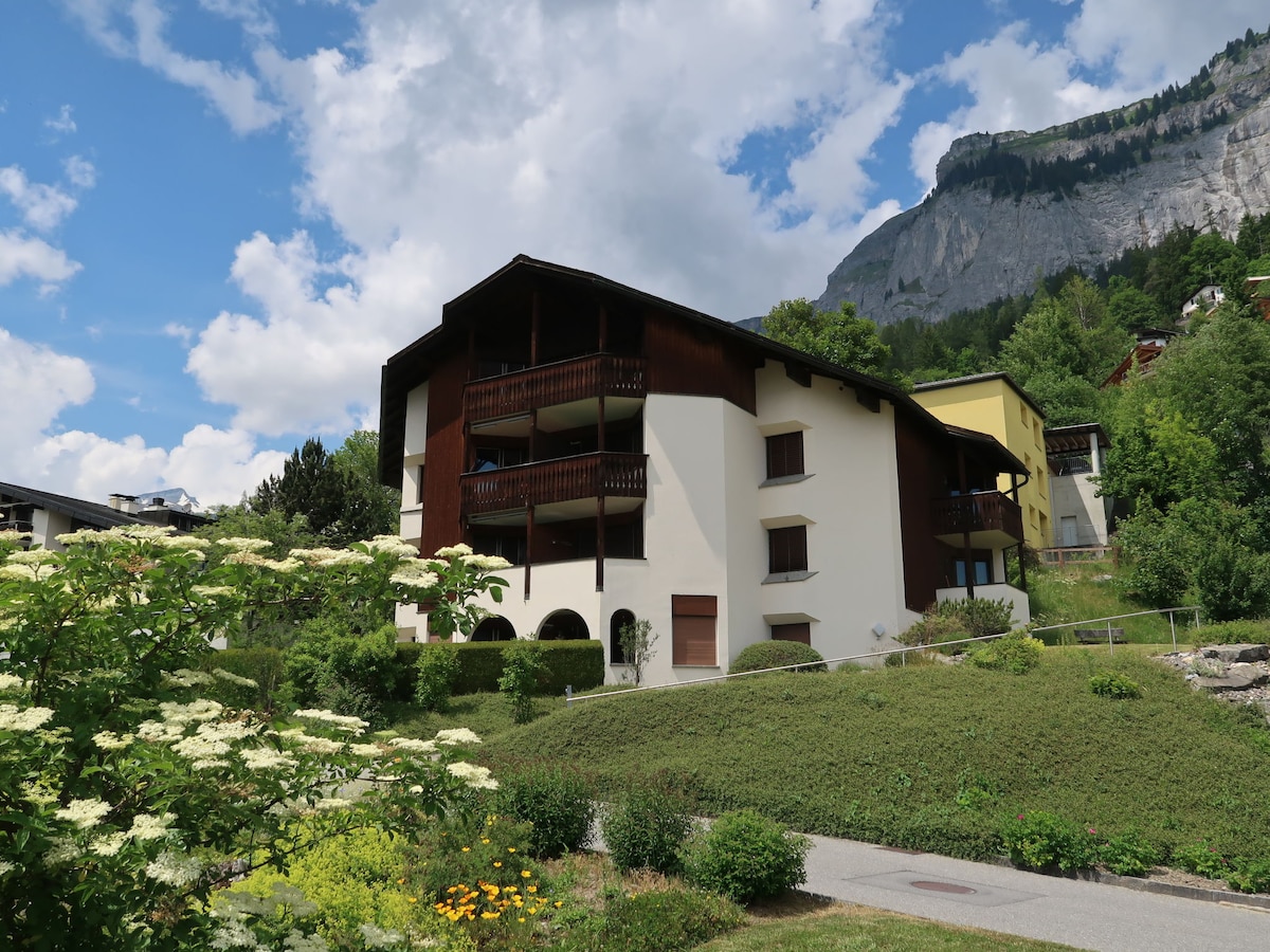 The exterior of the apartment building is shown, featuring a mix of wooden and stucco elements. Lush greenery surrounds the property, with flowering shrubs and a well-maintained garden. A backdrop of a mountain adds a scenic touch under a partly cloudy sky.
