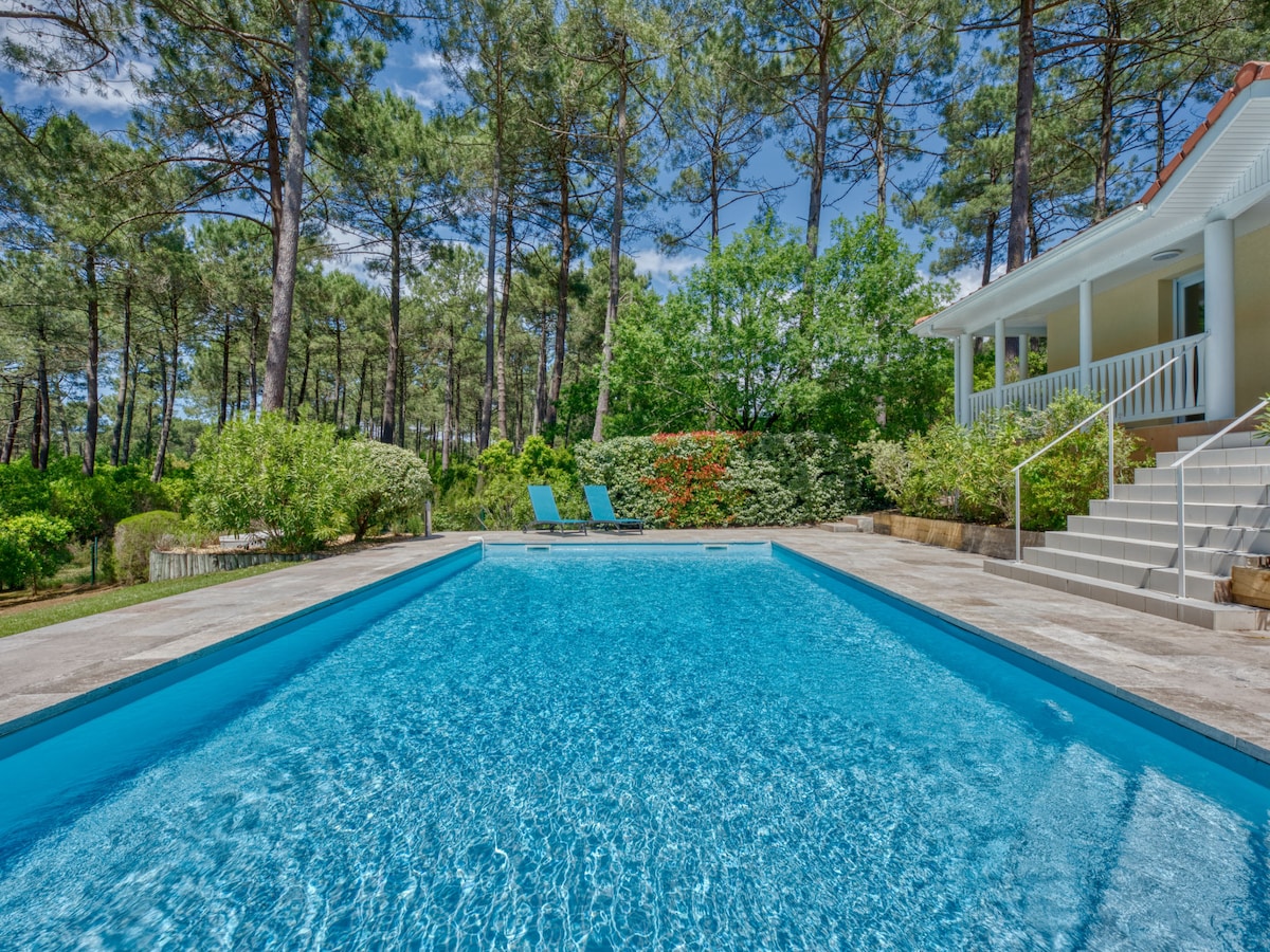 A clear swimming pool is set against a backdrop of lush greenery and tall trees. Two lounge chairs are positioned nearby on a sunlit patio. Steps lead from the pool area to a light-colored villa surrounded by well-maintained gardens.