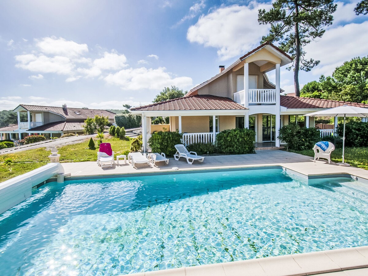 A spacious villa features a private pool, surrounded by sun loungers and a small garden area. The building showcases a two-level design with large windows and a balcony overlooking the pool. Clear skies and soft sunlight enhance the outdoor relaxation space, inviting guests to unwind.