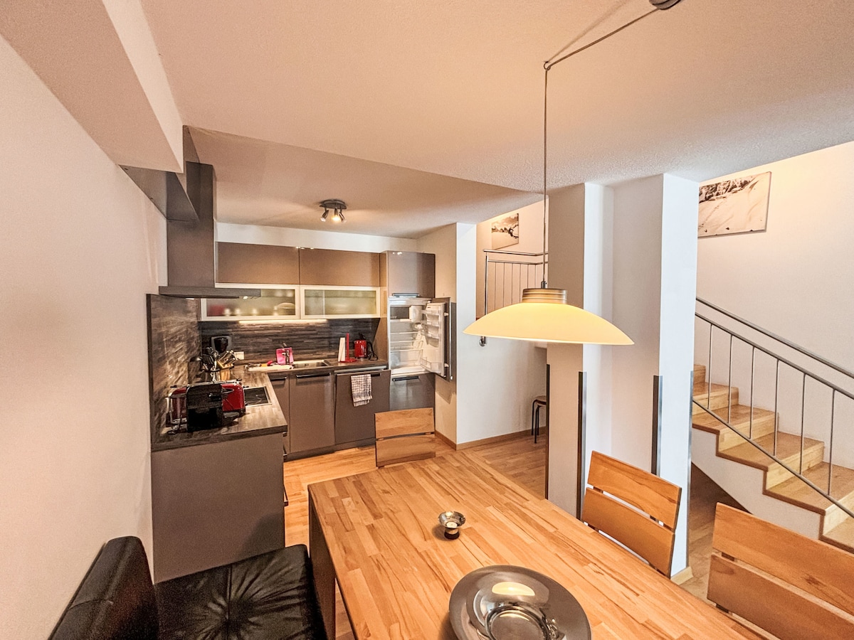 The open kitchen features modern appliances, including a refrigerator and an electric stove. A wooden dining table accommodates multiple chairs. Ample natural light illuminates the space, creating a functional area for cooking and dining. Stairs lead to an upper level, enhancing the layout.