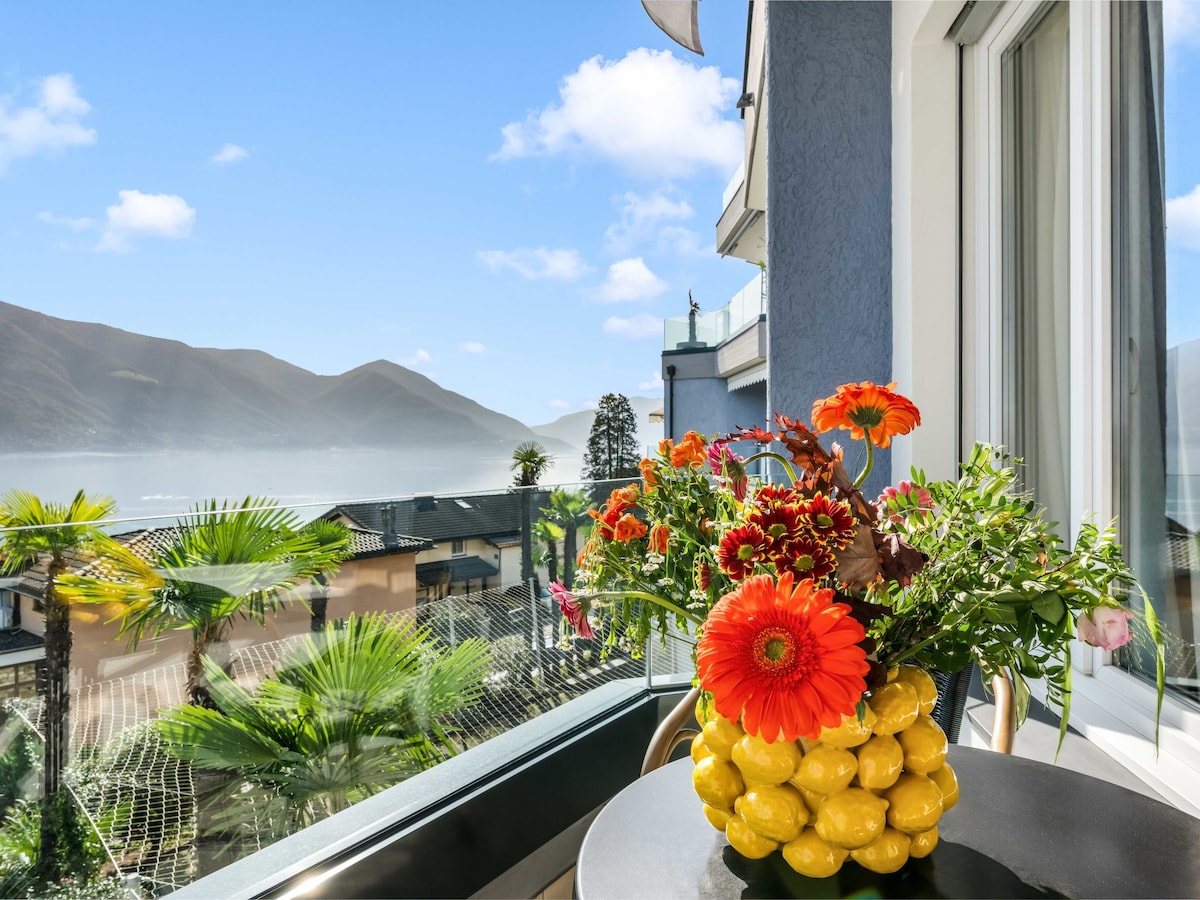 A balcony view is presented, featuring a vibrant arrangement of flowers and fruit. The colorful display includes orange gerbera daisies and yellow lemons. A landscape of mountains and palm trees is visible in the background, with soft morning light enhancing the serene ambiance.