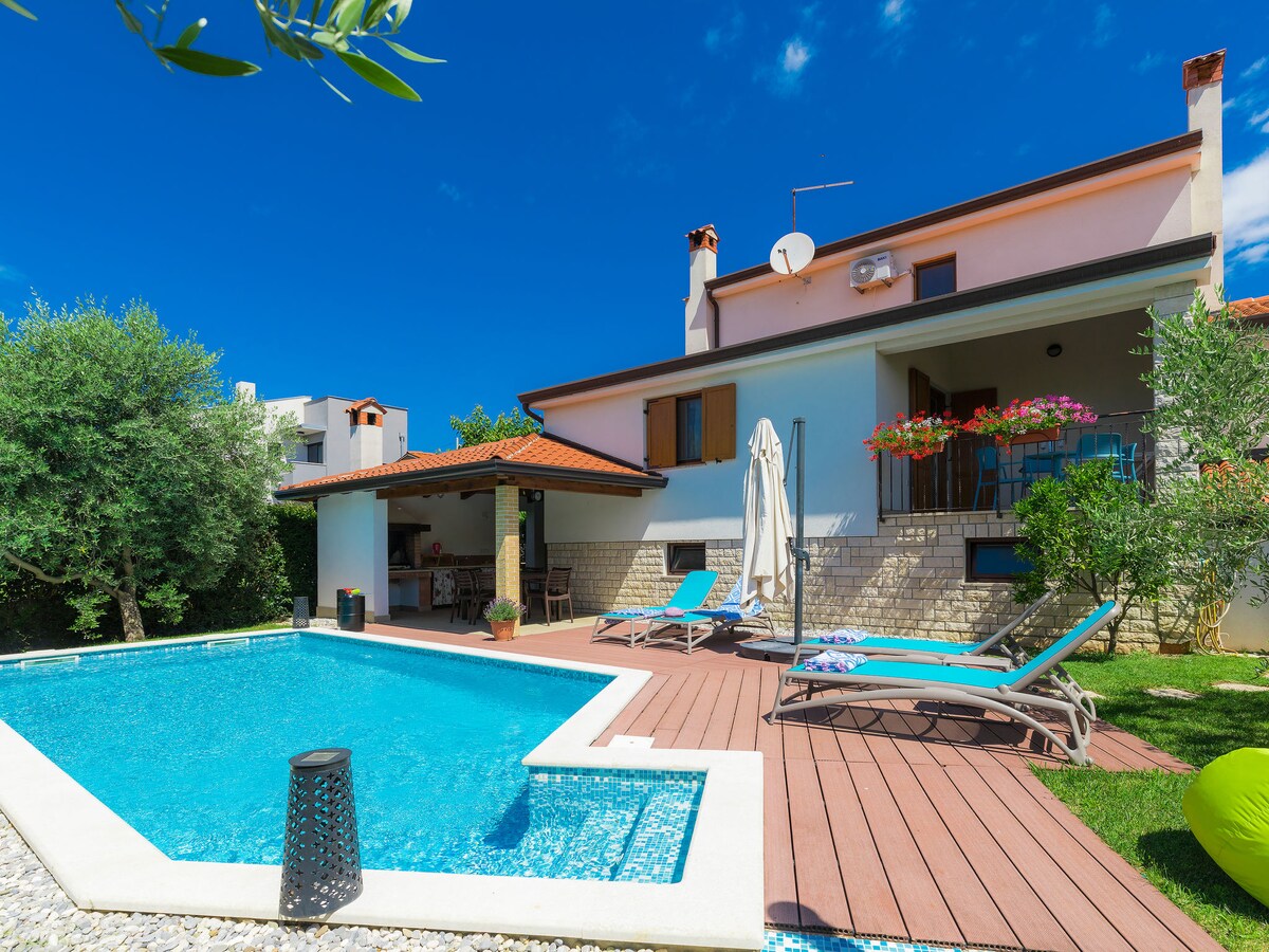 The exterior showcases a spacious garden with a private swimming pool, surrounded by a wooden deck and sun loungers. A covered terrace is visible, adorned with potted flowers, contributing to the vibrant atmosphere against a clear blue sky.