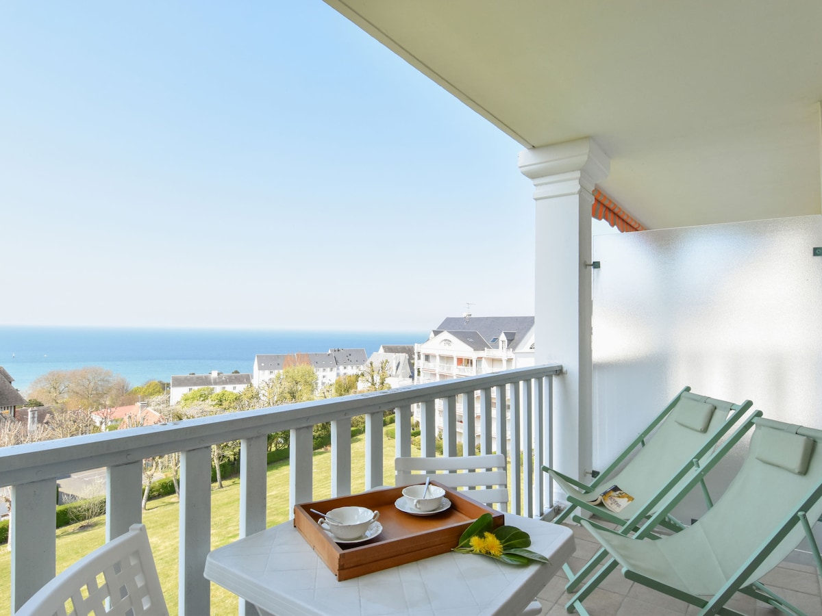 A spacious balcony features a table set with two plates and a flower, complemented by two lounge chairs. The view extends over lush greenery towards the sea, with a clear blue sky overhead. The balcony's railing provides an open and airy feel.