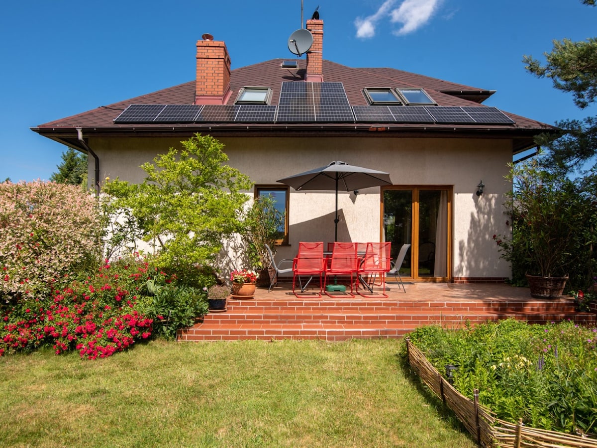 The exterior of a two-story house features a private terrace with red chairs and an umbrella, surrounded by a well-maintained garden. Solar panels are visible on the roof, and colorful flowers add charm to the landscape.