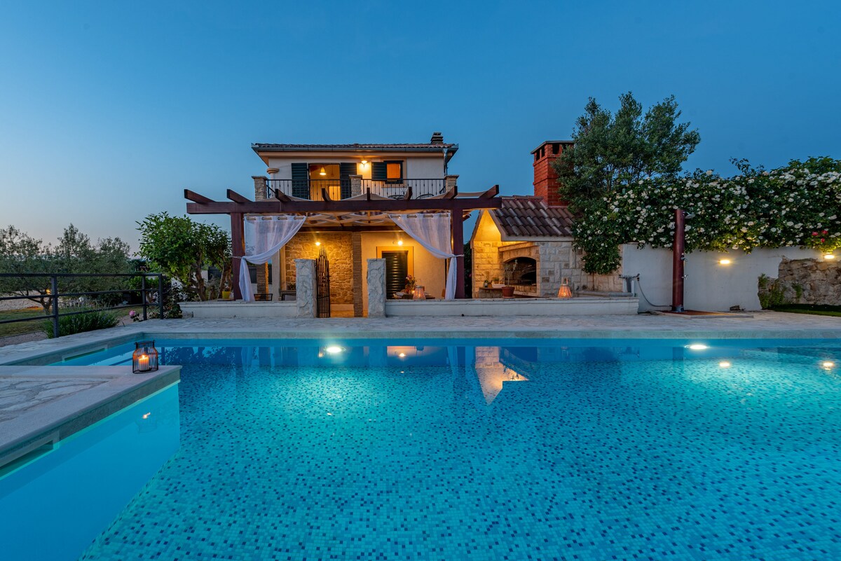 The villa's exterior is visible beside a large swimming pool reflecting the evening sky. A covered terrace features soft drapes and leads to the entrance. The rustic stone facade is complemented by lush greenery and built-in barbecue facilities, creating a serene outdoor space.
