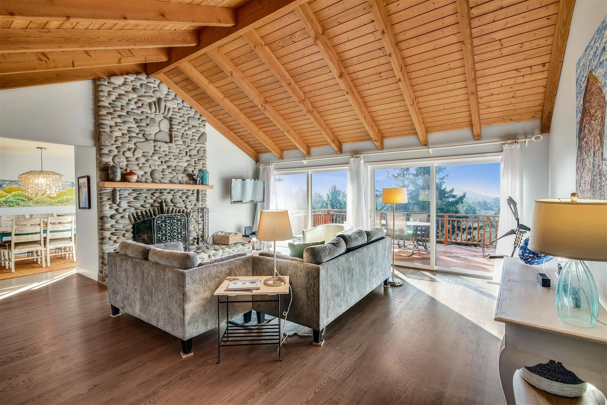 A spacious living area is highlighted by a high wooden ceiling and large windows that frame scenic views. A stone fireplace and comfortable seating create a cozy environment, complemented by a nearby deck access. Natural light enhances the warm tones of the furnishings.