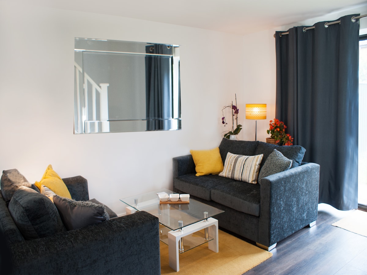 A modern living space features two dark sofas adorned with yellow cushions. A glass coffee table sits centrally, complemented by a soft area rug. A floor lamp emits warm light, and a mirrored accent reflects the room's layout, accompanied by a potted flowering plant.