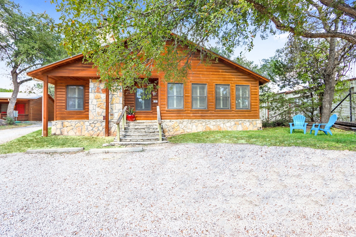 A welcoming log cabin features a stone and wooden exterior, complemented by a spacious front porch with steps leading to the entrance. Two bright blue chairs are positioned on the manicured lawn, providing an inviting outdoor seating area amidst a tree-lined setting.