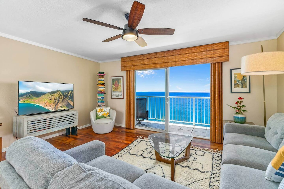 A spacious living area features a comfortable grey sectional sofa and a glass coffee table. A flat-screen television is positioned against a wall, while a large window showcases unobstructed ocean views and fills the room with natural light. Ceiling fan adds to the airy atmosphere.
