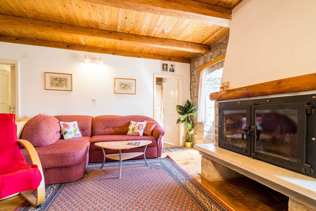 A cozy living area is presented with a plush red sofa and decorative cushions. A circular wooden table is situated in front of a stone fireplace. Natural light enters through the large window, illuminating the wooden ceiling and rustic stone walls.
