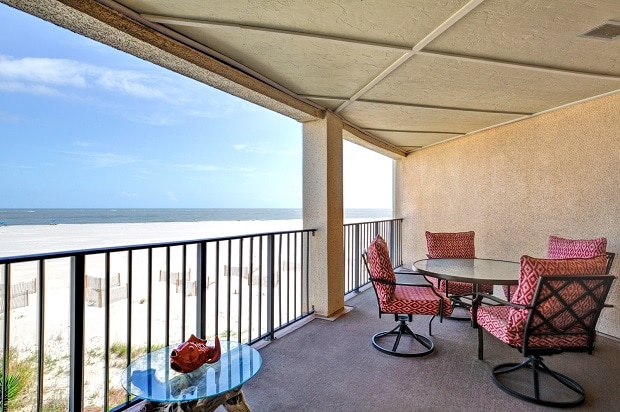 A spacious balcony features a round dining table surrounded by four cushioned chairs, offering a comfortable setting. The railing provides an unobstructed view of the beach and ocean beyond, where gentle waves meet the sand under a bright sky.