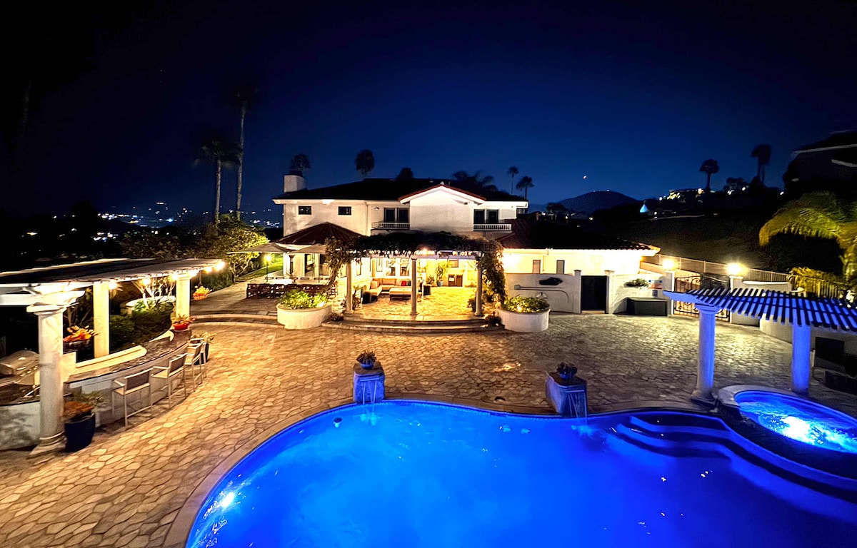 The villa is illuminated at night, showcasing a spacious outdoor area with a blue pool and stone patio. Surrounding seating arrangements create a welcoming atmosphere, while palm trees and distant city lights enhance the tranquil ambiance.