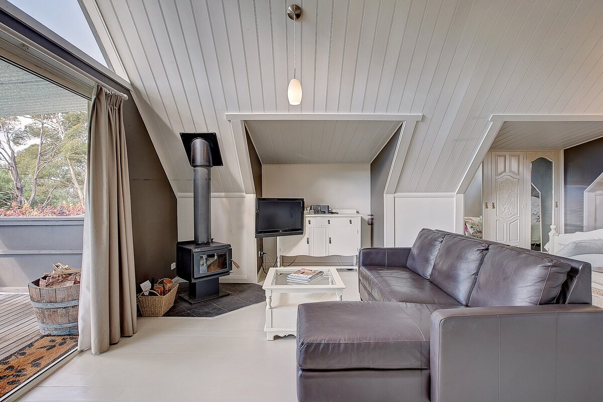 An inviting loft space features a leather sectional sofa facing a modern television and a compact fireplace. Natural light streams in through large windows adorned with flowing curtains, and a coffee table sits at the center, enhancing the relaxed atmosphere.