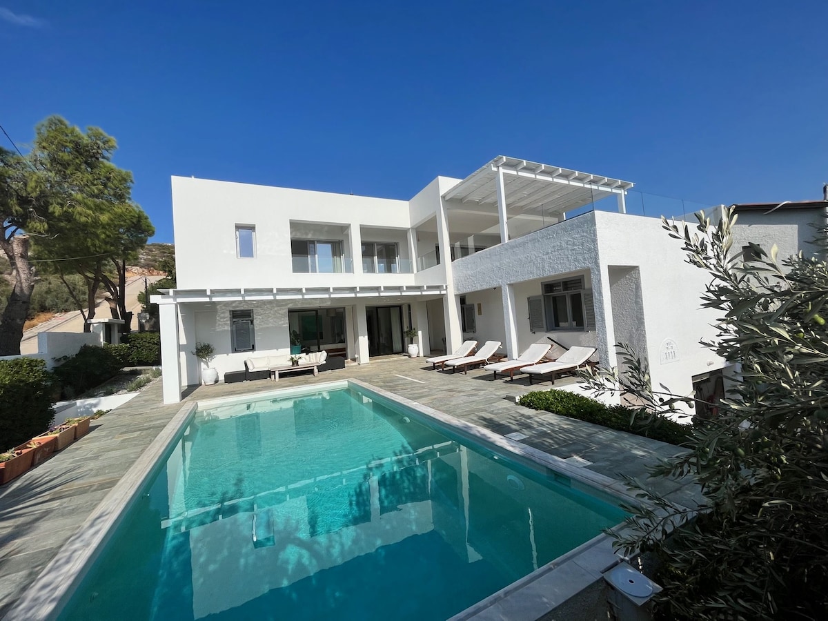 The modern villa features a sleek exterior with a private pool reflecting the clear blue sky. Loungers are arranged along the poolside, surrounded by manicured greenery and stone pathways. Spacious outdoor areas invite relaxation, while the two-story structure showcases expansive glass doors for natural light.