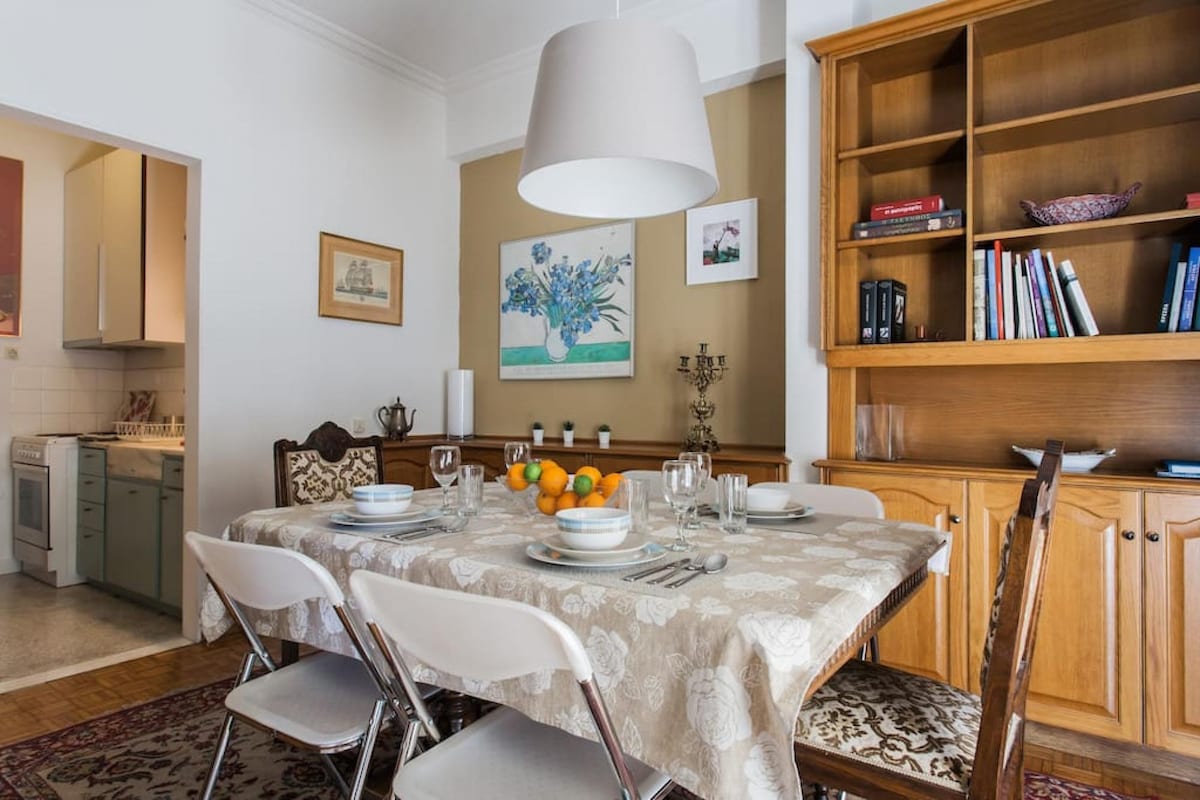 A dining area features a large table set with plates, cutlery, and glassware, surrounded by chairs. A bowl of oranges adds a pop of color. Walls are adorned with artwork and shelves are filled with books, while a light fixture hangs above, creating a functional and welcoming space.