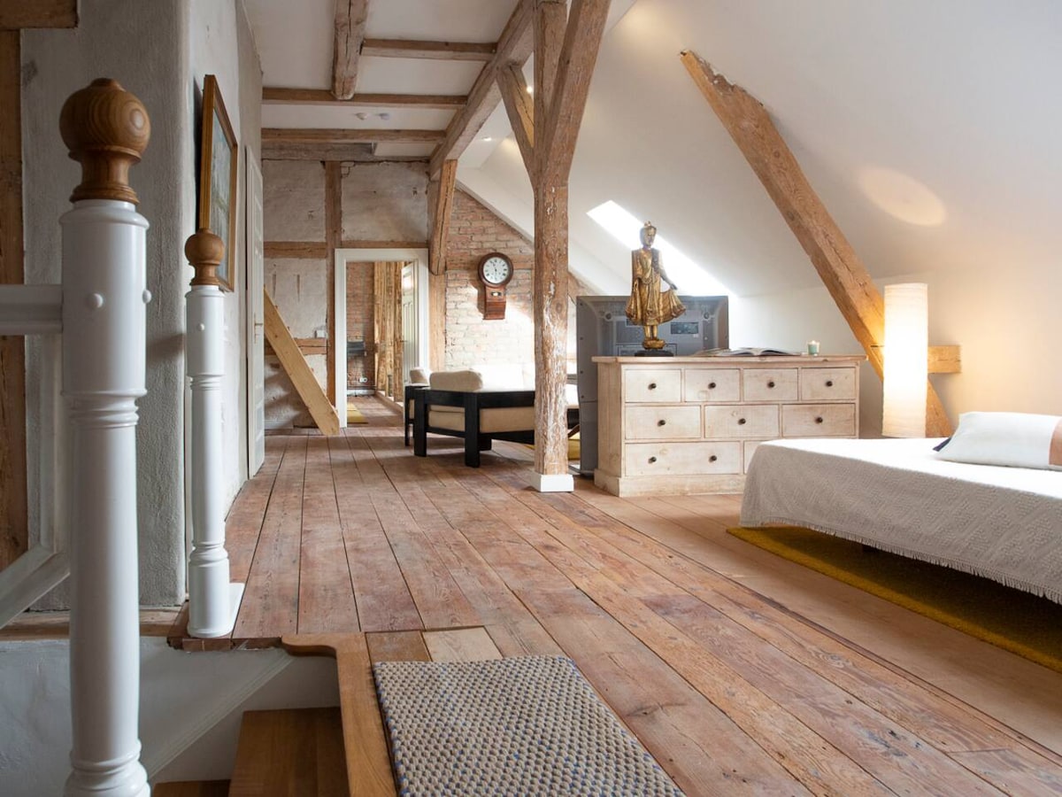 An airy loft space features exposed wooden beams and a warm wooden floor. A spacious bed is positioned against a slanted wall, with a cozy throw draped over it. A stylish dresser and seating area with a dark sofa are present, creating a comfortable and inviting atmosphere.
