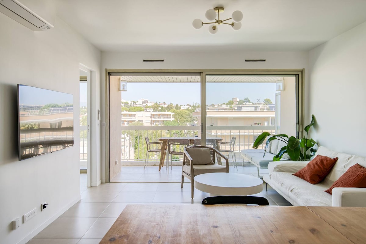 A spacious living area is presented with a comfortable sofa adorned with decorative pillows. A large window provides access to natural light and offers views of the terrace. A dining table is visible, accompanied by additional seating on the terrace, creating an inviting environment.